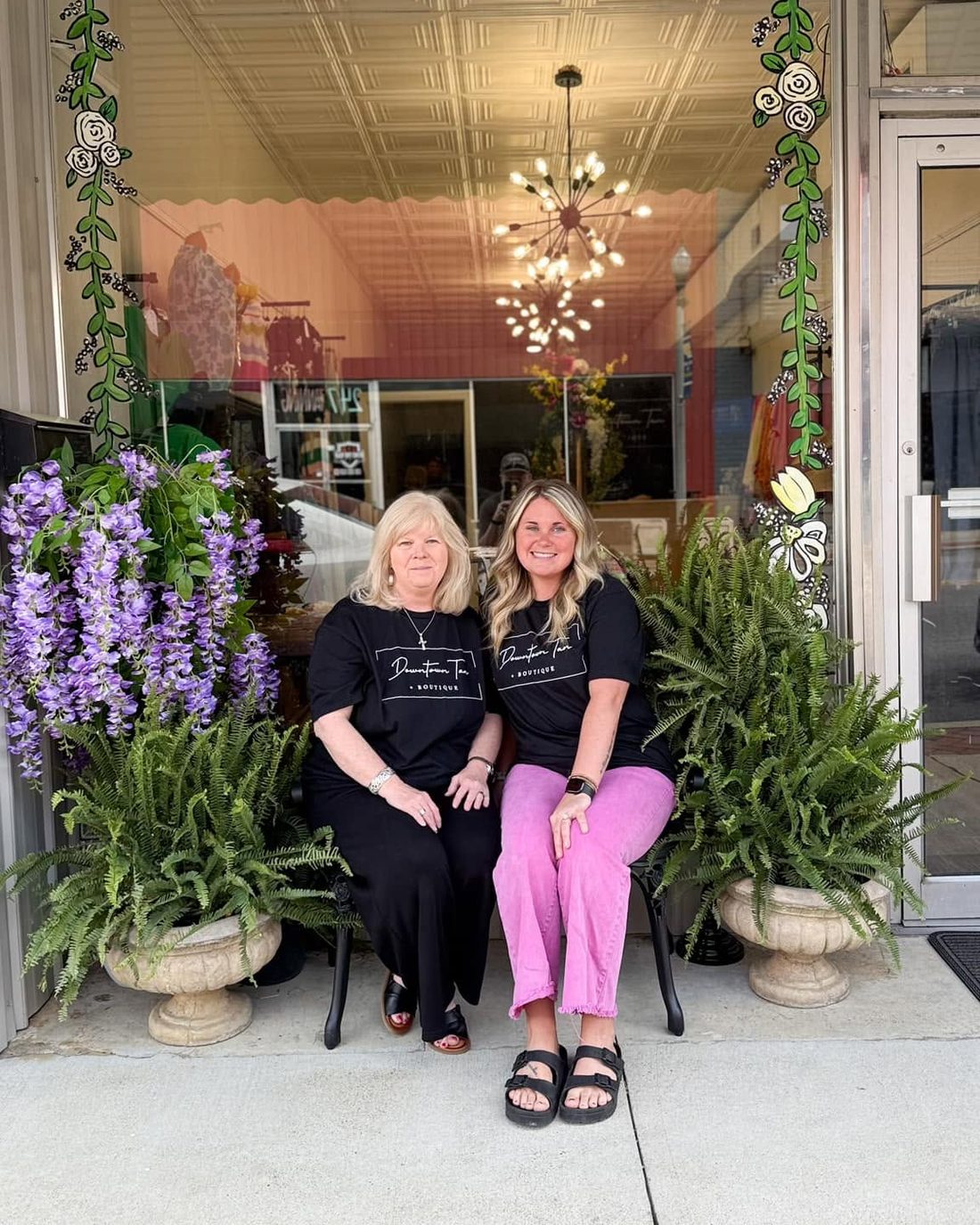 The two owners of Downtown Tan + Boutique seated in front of the shop between ferns and hanging wisteria